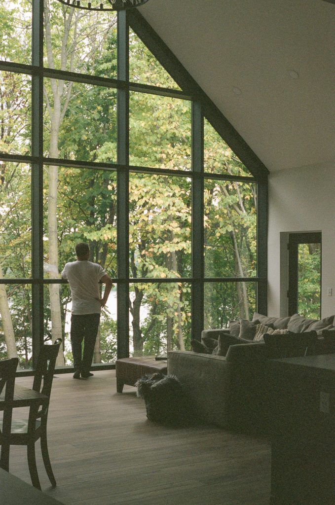 A man looks out floor to ceiling windows overlooking a lake in this residential interior photograph made on film.